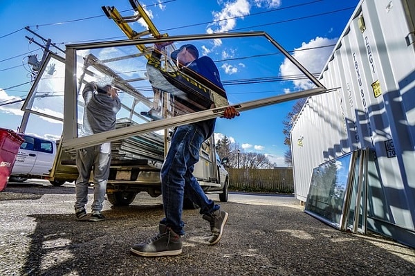 Two workers in jeans and caps carrying a large double-glazed window with its frame, in front of a service truck and containers.